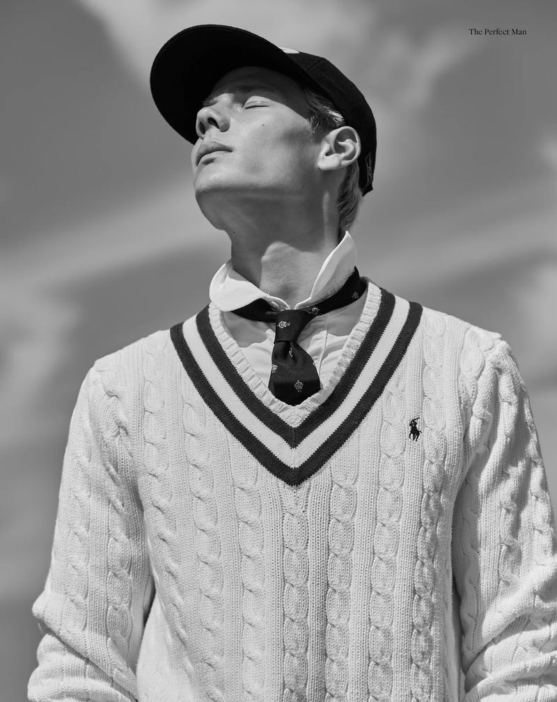 Model Blue River Wild wearing a white cable knit cricket sweater, tie, and cap looking up against an outdoor sky in black and white.