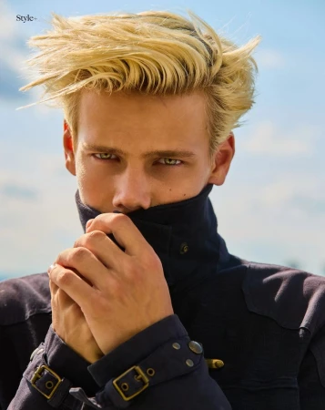 Model Blue River Wild looking at the camera while covering his lower face with the high collar of a navy jacket outdoors.