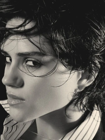 Black and white close-up portrait of Sasha Calle with wet-look short dark hair wearing a striped collared shirt against a black background.