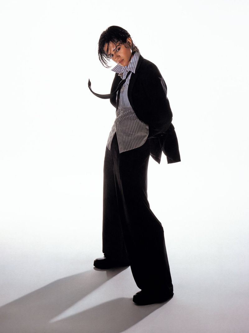 Sasha Calle posing in an oversized black suit, striped button-up shirt, and loose black necktie against a bright white seamless background.