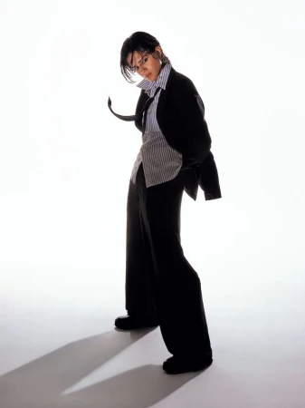 Sasha Calle posing in an oversized black suit, striped button-up shirt, and loose black necktie against a bright white seamless background.