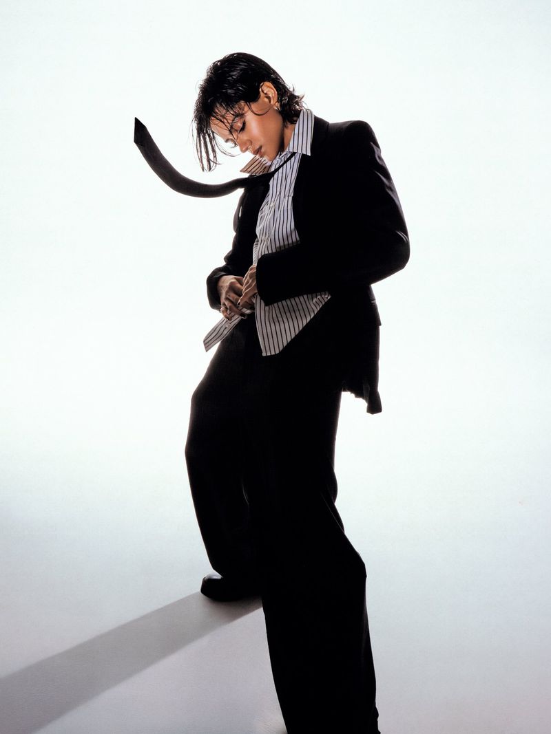 Sasha Calle posing in an oversized black suit, striped shirt, and floating black necktie against a bright white background.