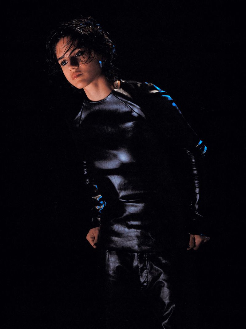 Sasha Calle posing in a glossy black long-sleeve top and trousers against a dark background in low-key lighting.