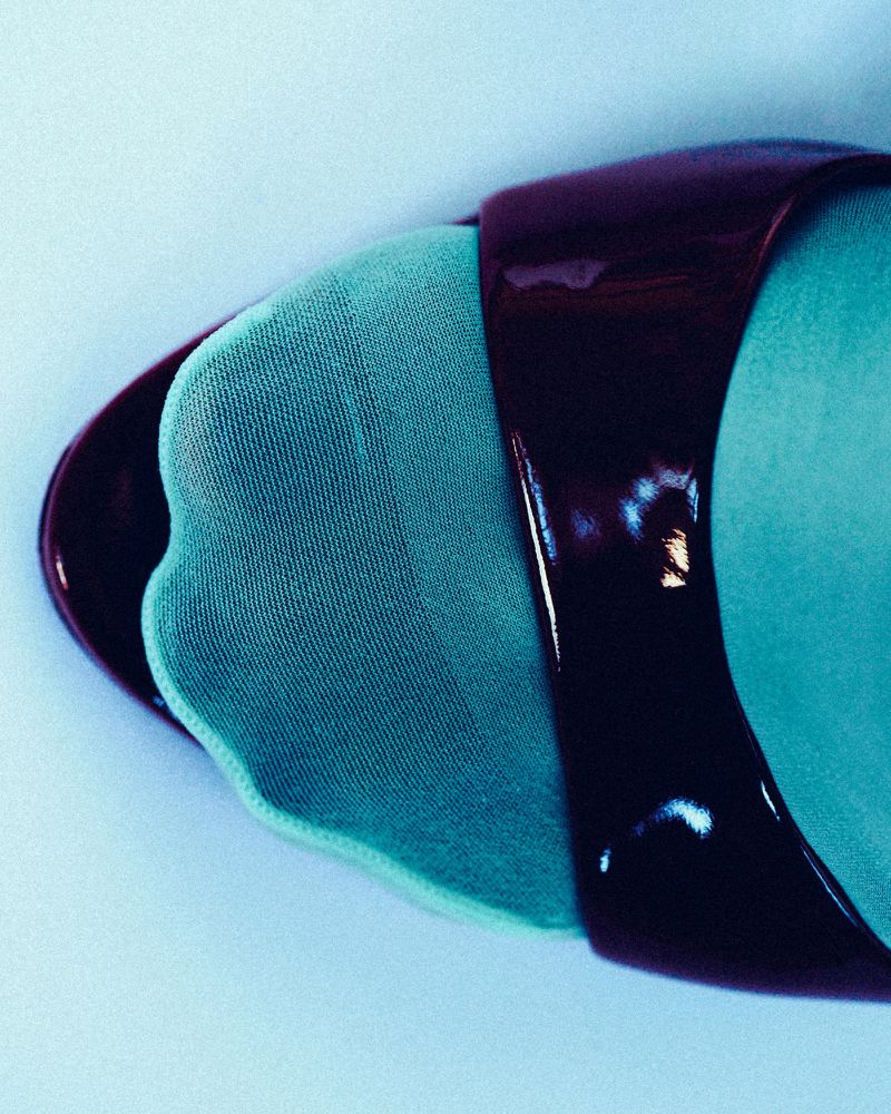 Close-up of a foot in bright turquoise stockings wearing a glossy dark burgundy patent leather open-toe strap shoe.