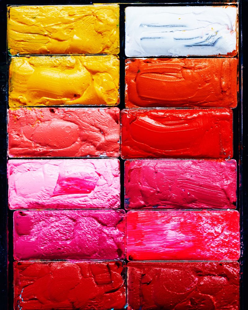 Close-up overhead view of a makeup palette with rectangular pans of thickly smeared, brightly colored cream pigments in yellows, pinks, and reds.
