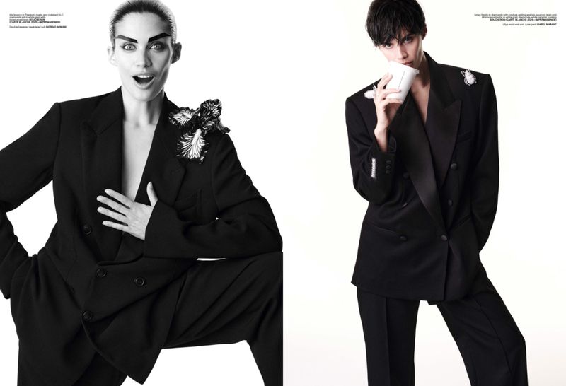 Editorial fashion shot of two models in black suits against a white backdrop, one drinking from a white cup, showcasing bold makeup.