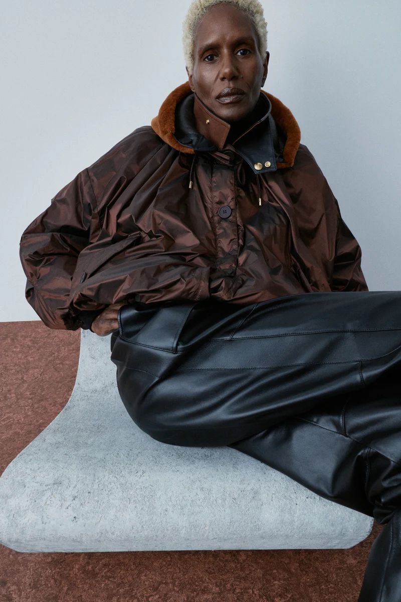 Model with short blonde hair seated on a curved stone bench wearing a voluminous bronze jacket and black leather pants.