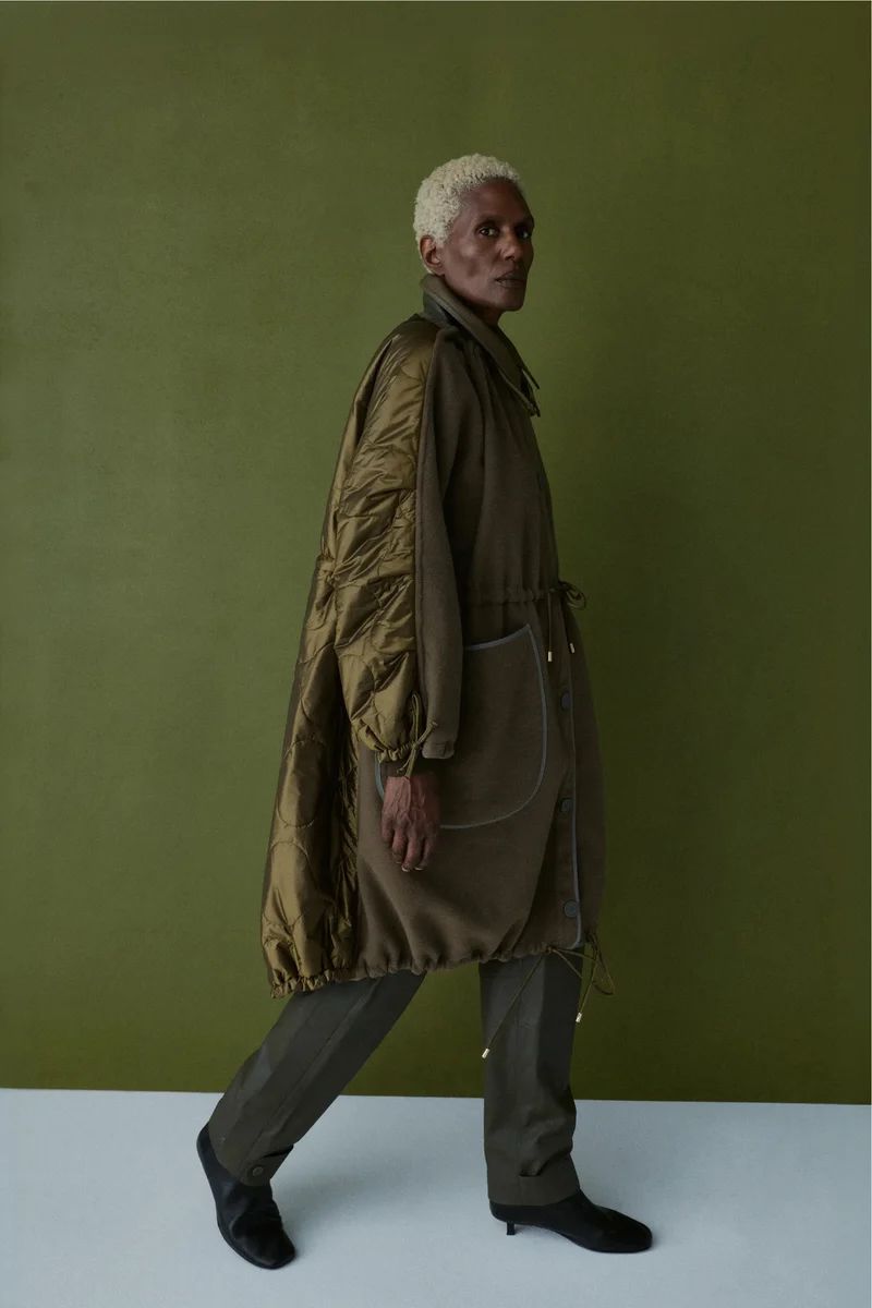 Model with short blonde hair walking in profile wearing an olive green quilted coat and trousers against a green studio backdrop.