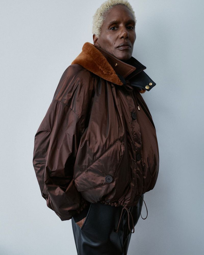 Model with short blonde hair wearing an oversized brown jacket with a fur collar and black trousers against a neutral background.