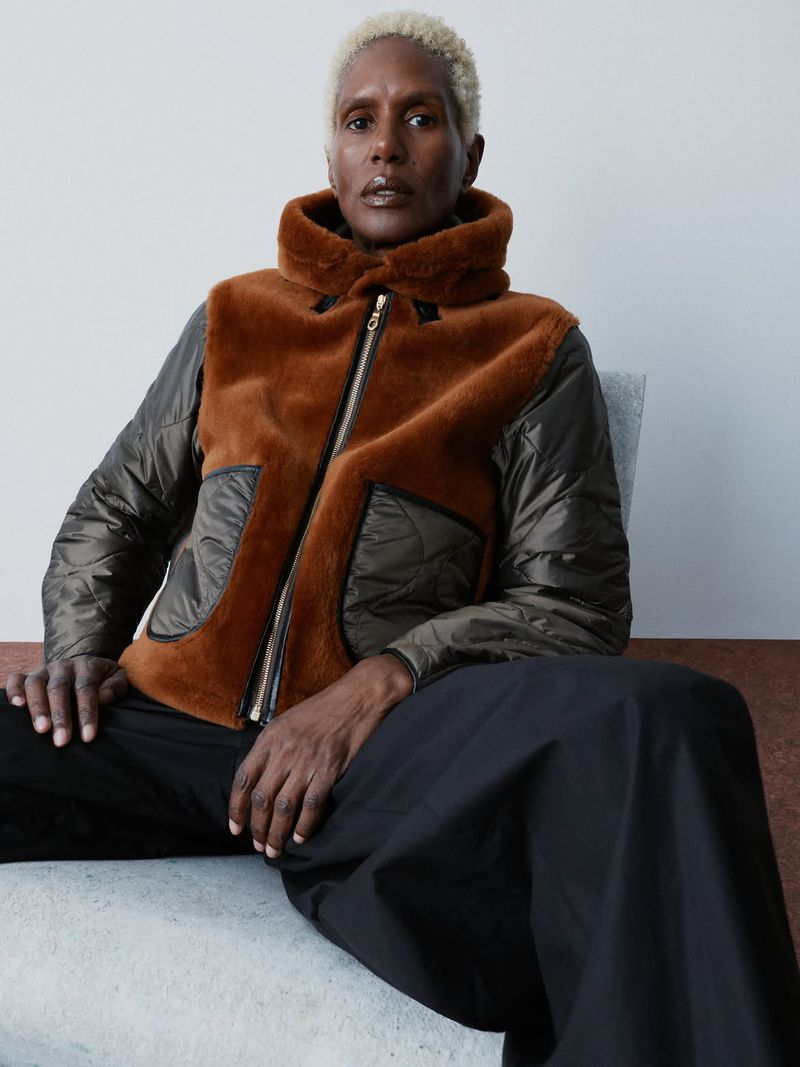 Model seated wearing a brown shearling and olive quilted jacket with wide black trousers against a pale studio backdrop.