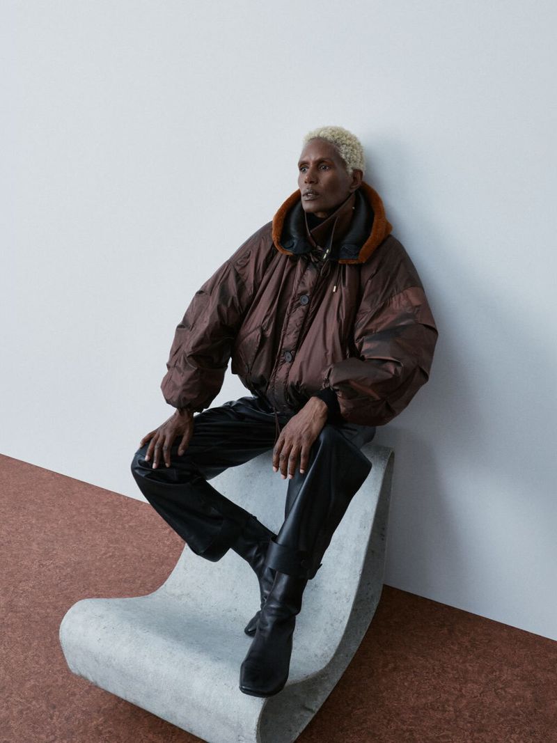 Model Amanda Parker in an oversized brown hooded jacket and black pants seated on a curved concrete bench against a white wall.