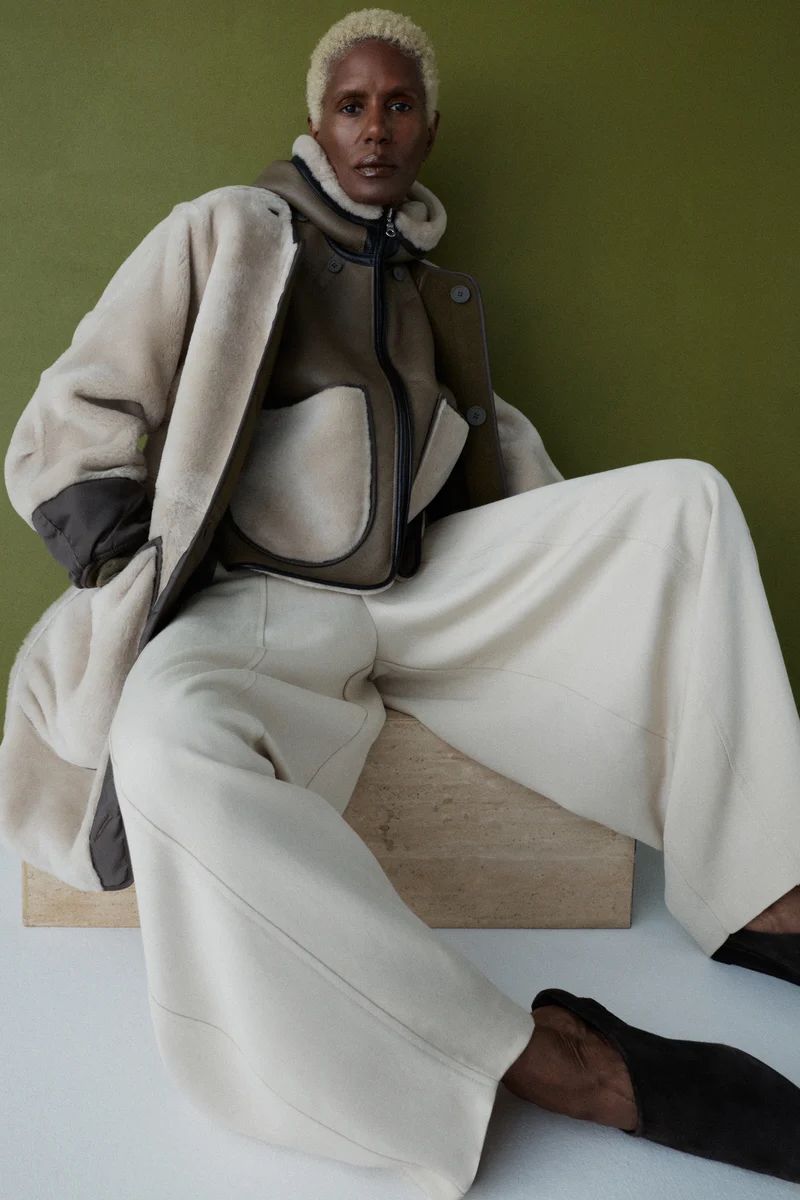Model Amanda Parker seated in layered cream shearling outerwear and wide-leg trousers against an olive green backdrop.