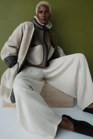 Model Amanda Parker seated in layered cream shearling outerwear and wide-leg trousers against an olive green backdrop.