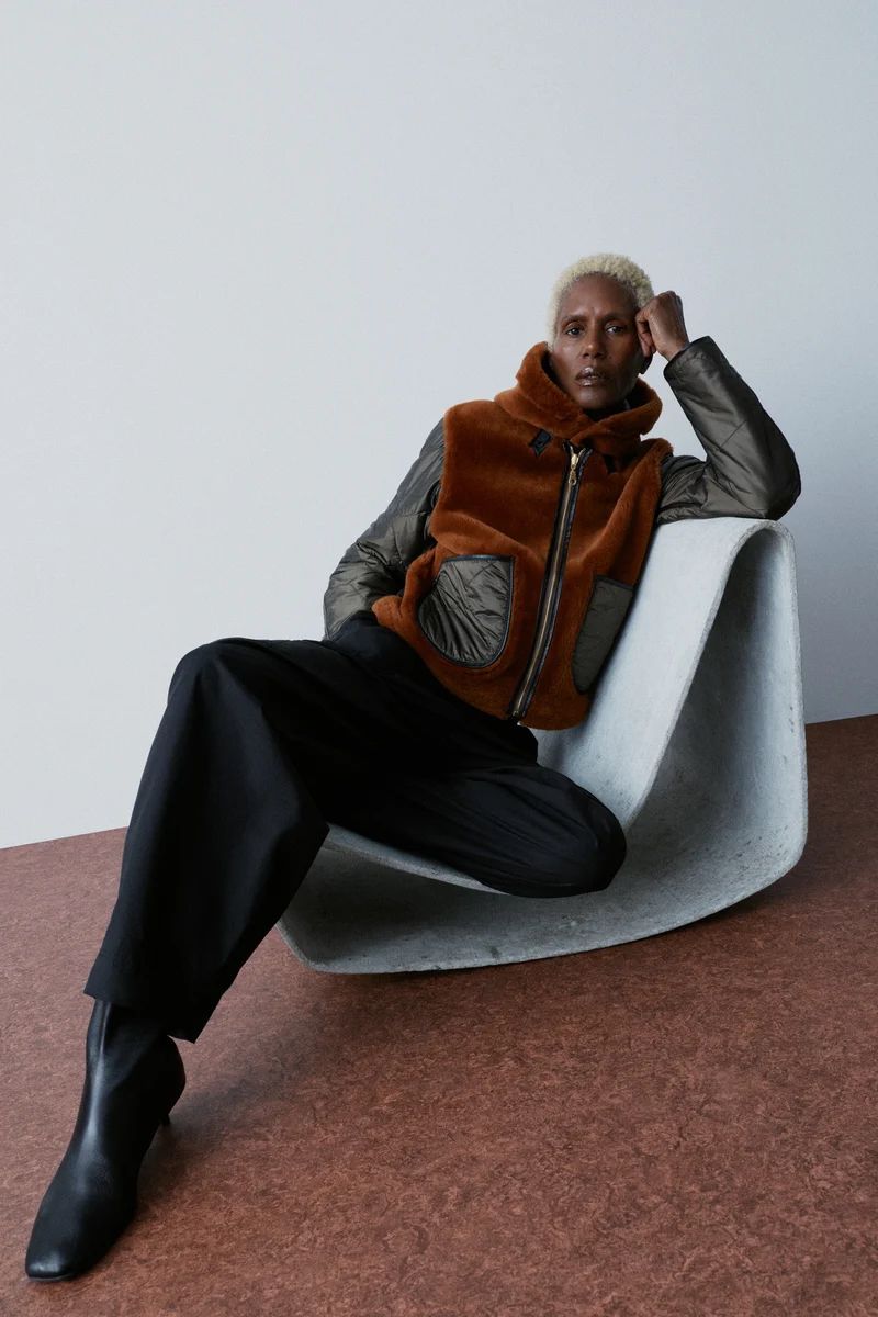 Model Amanda Parker seated in a curved concrete chair wearing a brown shearling and quilted jacket with black pants and boots.