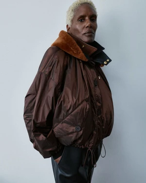 Amanda Parker wearing a voluminous brown bomber jacket with a fur-trimmed collar against a pale studio backdrop.
