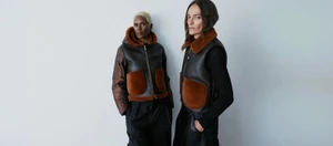 Two models wearing black leather and brown shearling outerwear posing together against a plain light studio backdrop.