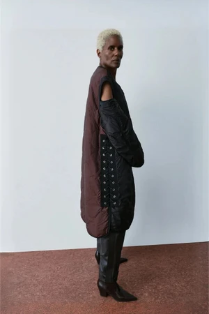 Model wearing a chocolate brown and black quilted lace-up tunic coat with black leather boots against a neutral studio backdrop.