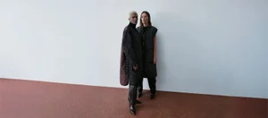 Two models standing together wearing black quilted outerwear against a white wall and reddish-brown floor.