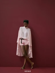 Model Anipha Umufite wearing a light pink draped cape top and matching shorts with a black belt against a dark burgundy background.