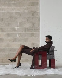 Model Anipha Umufite in a dark brown high-collar outfit seated on a sculptural armchair against a cinder block wall.