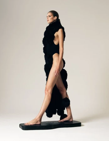 Barefoot model in a high-cut black bodysuit and textured black sculptural boa posing on a plinth against a white studio background.