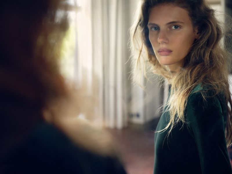 Female model wearing a dark green velvet long-sleeve dress looks back over her shoulder in a sunlit interior room.