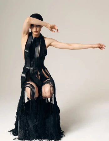 Model crouching in a black sculptural cutout dress with floor-length fringe against a white studio background.