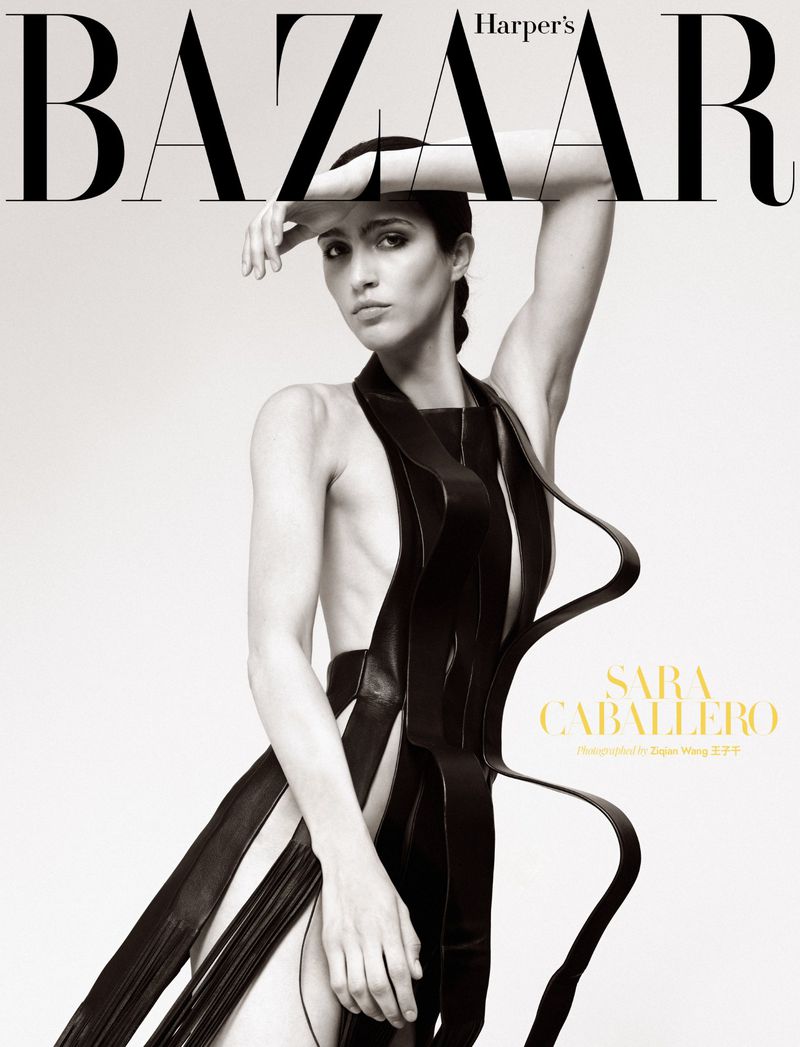 Model Sara Caballero posing with arm raised in a sculptural black leather ribbon garment against a seamless white studio background.