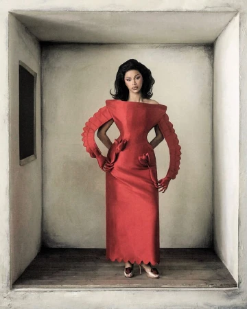 Cardi B posing in a sculptural red gown with pleated arm structures inside a neutral alcove set.