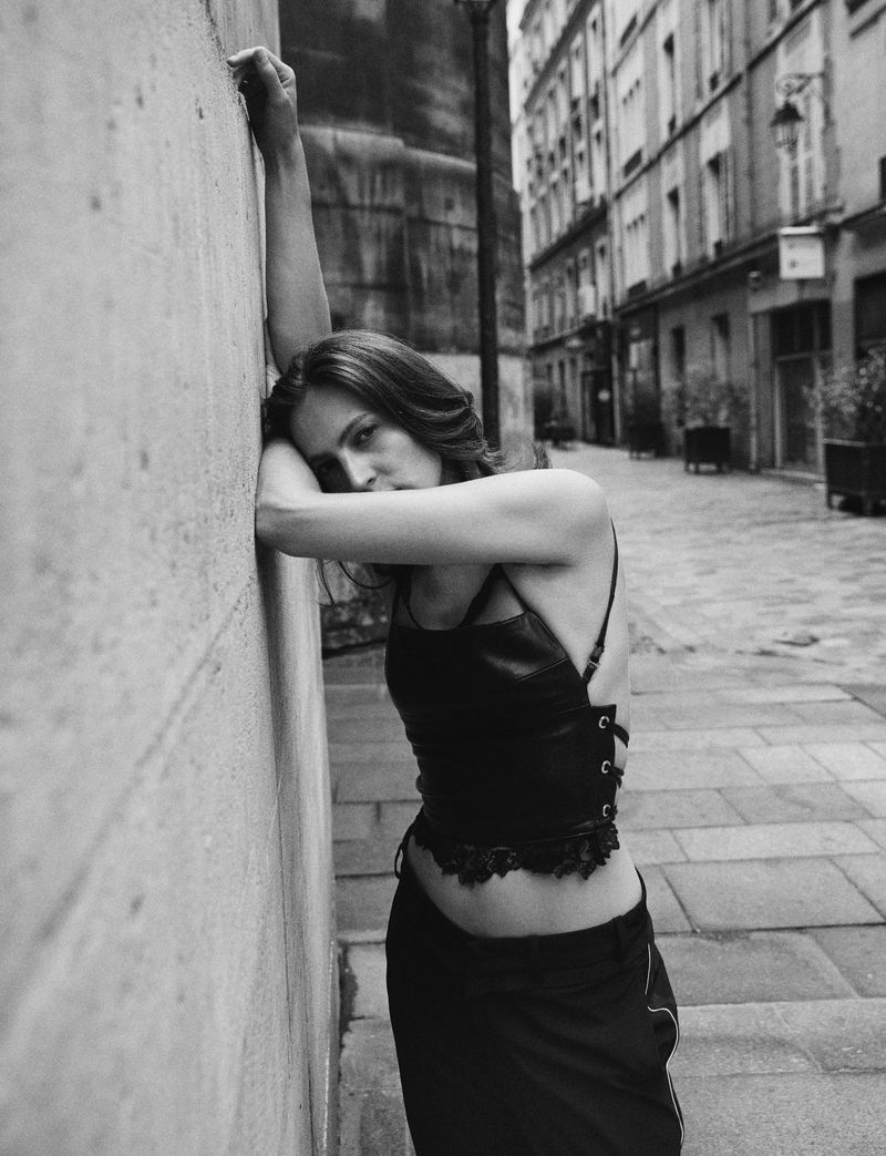 Black and white portrait of a slender adult woman in an alley, leaning against a concrete wall wearing a dark top and skirt.