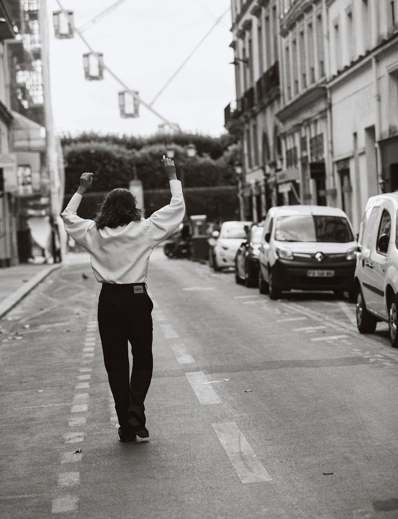 Back view of person in white shirt and black pants walking down a city street with arms raised in celebration in black and white.