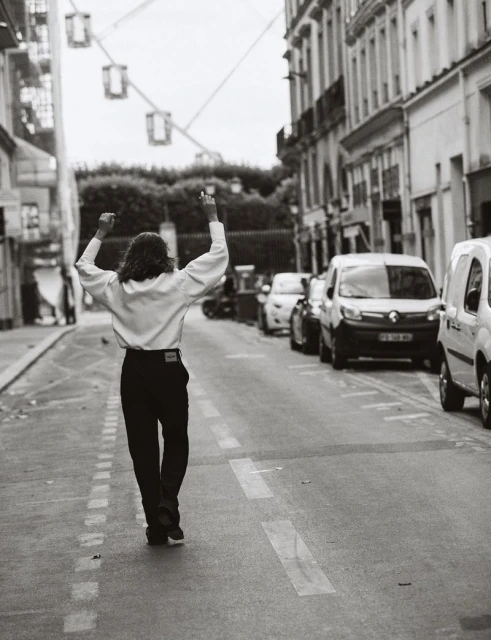 Back view of person in white shirt and black pants walking down a city street with arms raised in celebration in black and white.