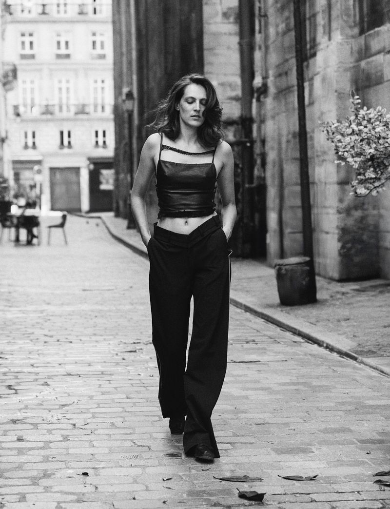 Monochrome full shot of a woman in a sleeveless crop top and slacks walking on a stone road flanked by buildings in a city.