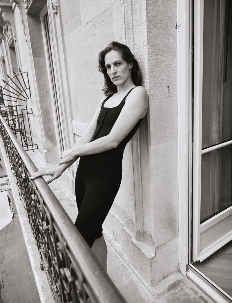 Adult in a black bodysuit stands on a balcony with an ornate metal railing, posing against a stone exterior in a black and white photograph.