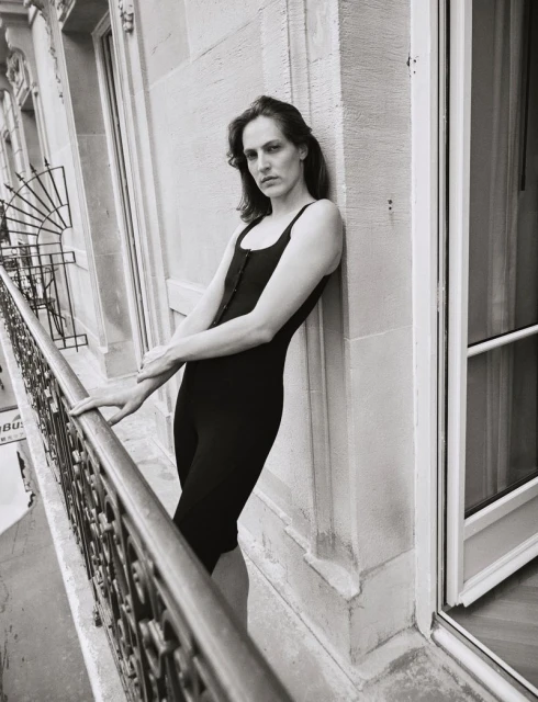 Adult in a black bodysuit stands on a balcony with an ornate metal railing, posing against a stone exterior in a black and white photograph.