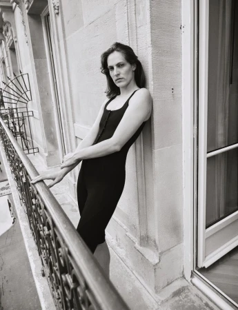 Adult in a black bodysuit stands on a balcony with an ornate metal railing, posing against a stone exterior in a black and white photograph.