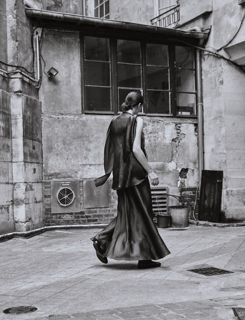 Monochrome image of a woman in a flowing gown walking in an urban courtyard, showcasing fashion and architecture in black and white tones.