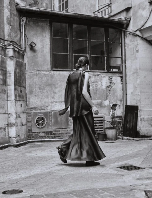 Monochrome image of a woman in a flowing gown walking in an urban courtyard, showcasing fashion and architecture in black and white tones.
