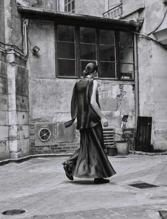 Monochrome image of a woman in a flowing gown walking in an urban courtyard, showcasing fashion and architecture in black and white tones.
