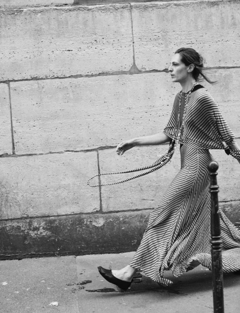 A slender adult female model walks on a sidewalk in a long, striped dress, posing in a black and white fashion photograph. 