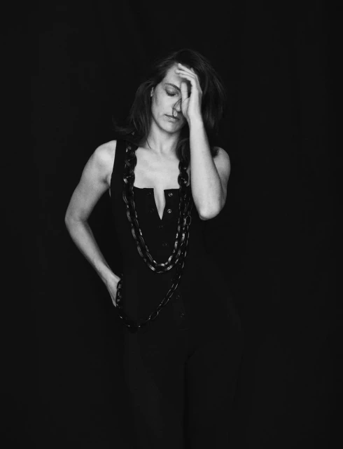 Monochrome portrait of a woman with a hand to her forehead wearing a dark sleeveless top and large beaded necklace against a black backdrop.