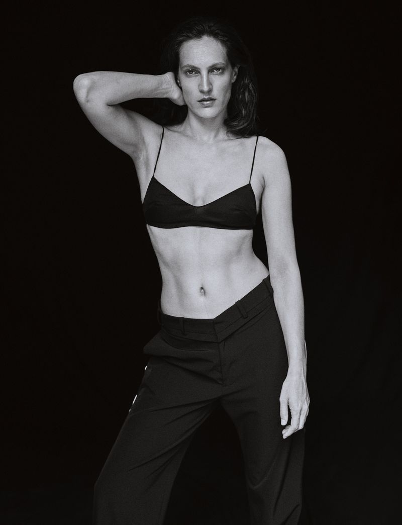Black and white studio portrait of a woman in a bra top and slacks against a dark backdrop, looking directly at the camera.