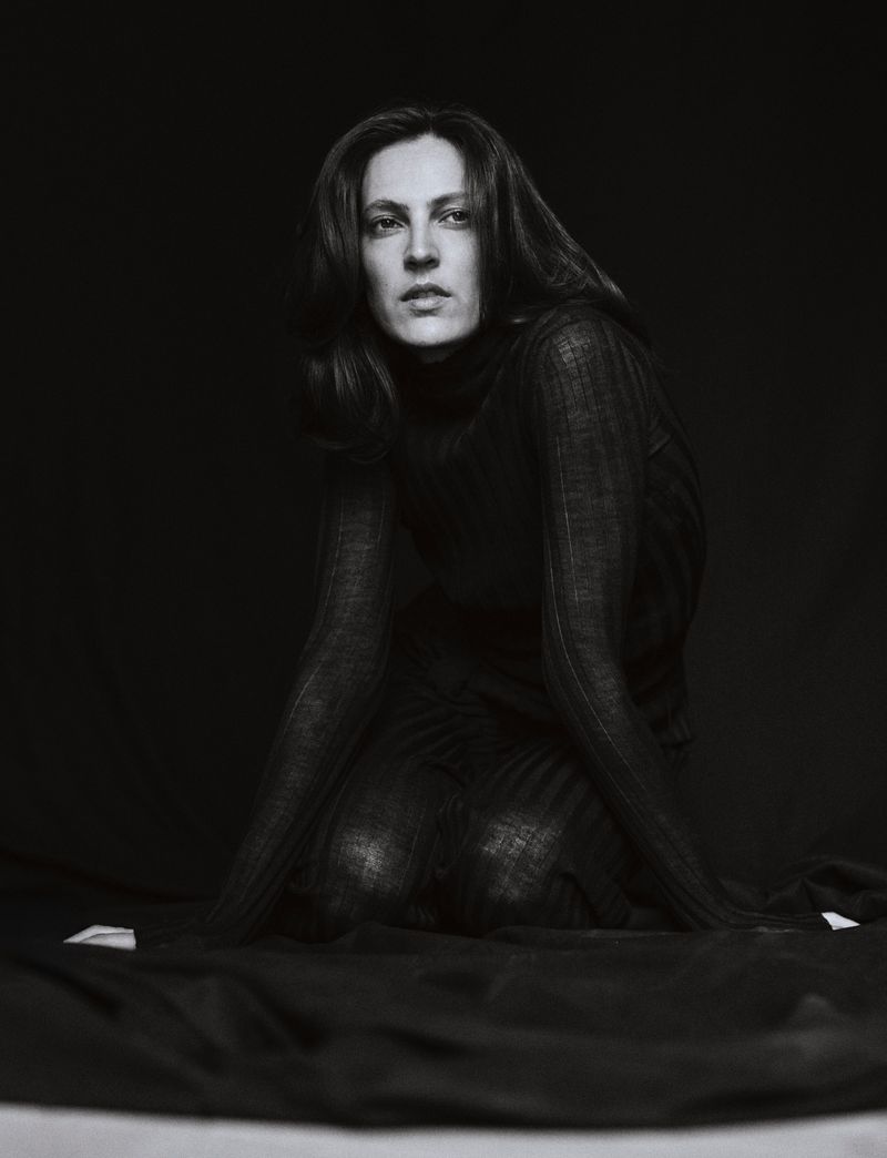 Monochrome portrait of a woman with long dark hair kneeling in a dark turtleneck sweater in front of a draped black fabric backdrop.