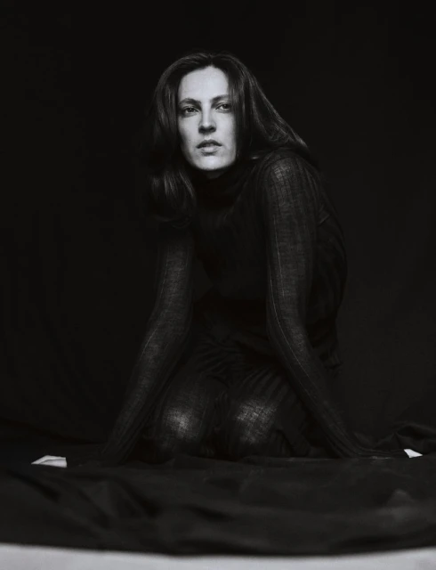 Monochrome portrait of a woman with long dark hair kneeling in a dark turtleneck sweater in front of a draped black fabric backdrop.