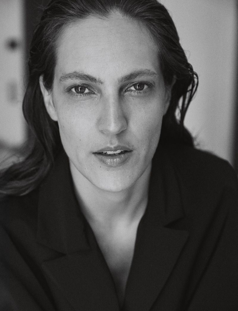 Headshot portrait of an adult woman with fair skin and dark hair wearing a black jacket against an indoor background in black and white.