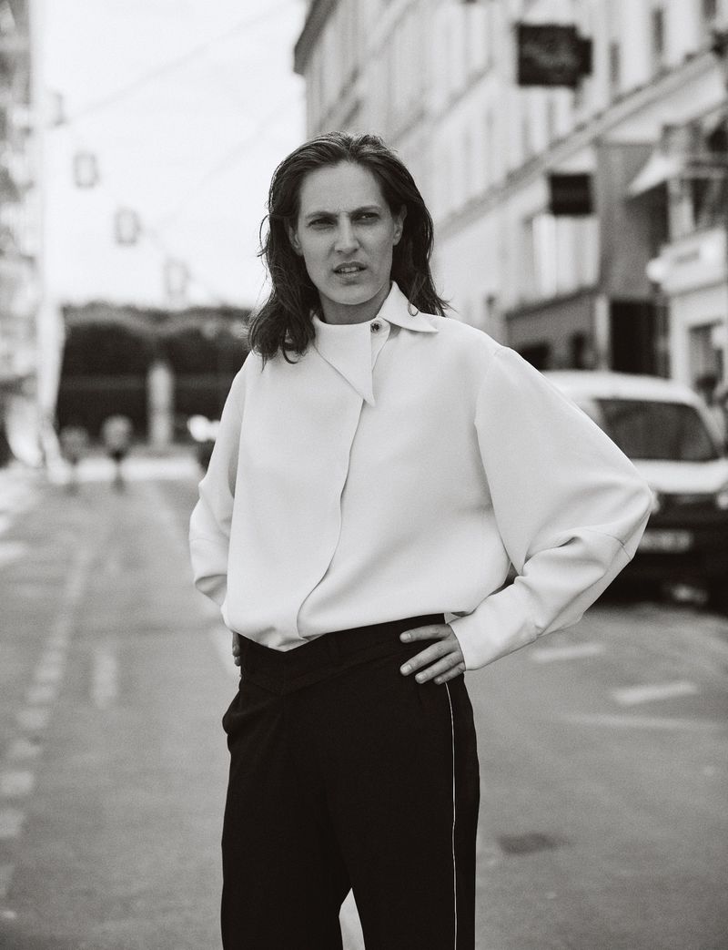 Black and white portrait of an adult woman standing on a city street, wearing a light-colored top and dark pants, looking at the camera.