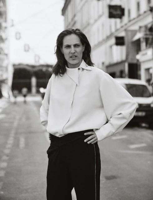 Black and white portrait of an adult woman standing on a city street, wearing a light-colored top and dark pants, looking at the camera.