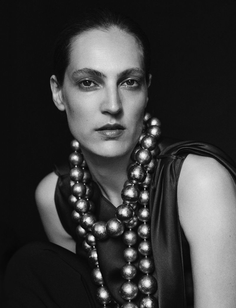 Monochrome portrait of an adult female model wearing a dark top and large, spherical beaded necklaces against a dark background.