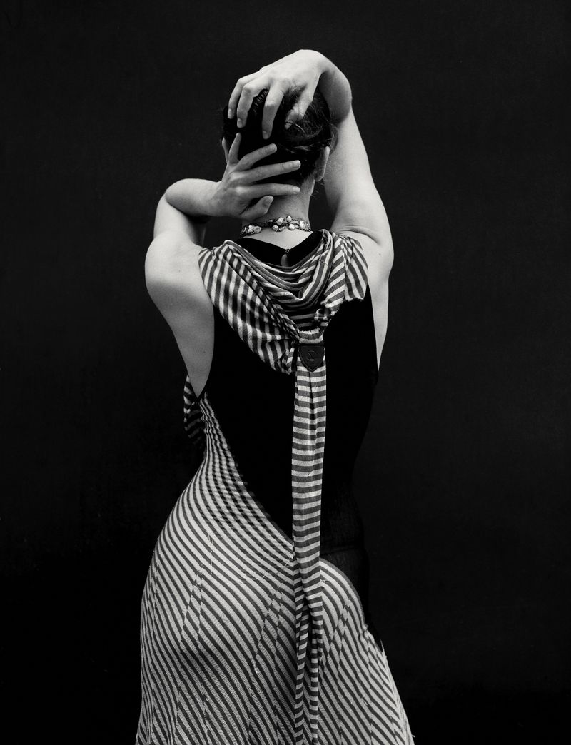 Elegant, black and white studio portrait of the back of a fair-skinned adult female wearing a striped dress, hands behind head.
