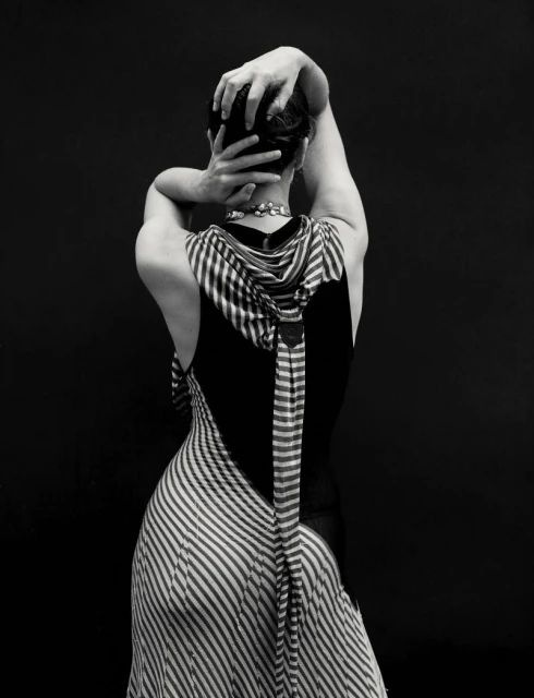 Elegant, black and white studio portrait of the back of a fair-skinned adult female wearing a striped dress, hands behind head.